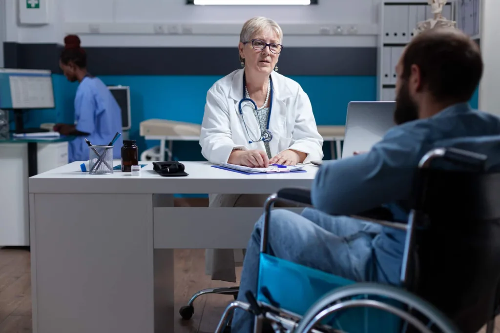 Un medico donna alla scrivania e, di fronte a lei e di spalle, un paziente su una sedia a rotelle.