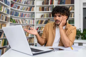 Uomo latino alla scrivania davanti a un portatile con espressione sorpresa e una mano che indica il monitor.